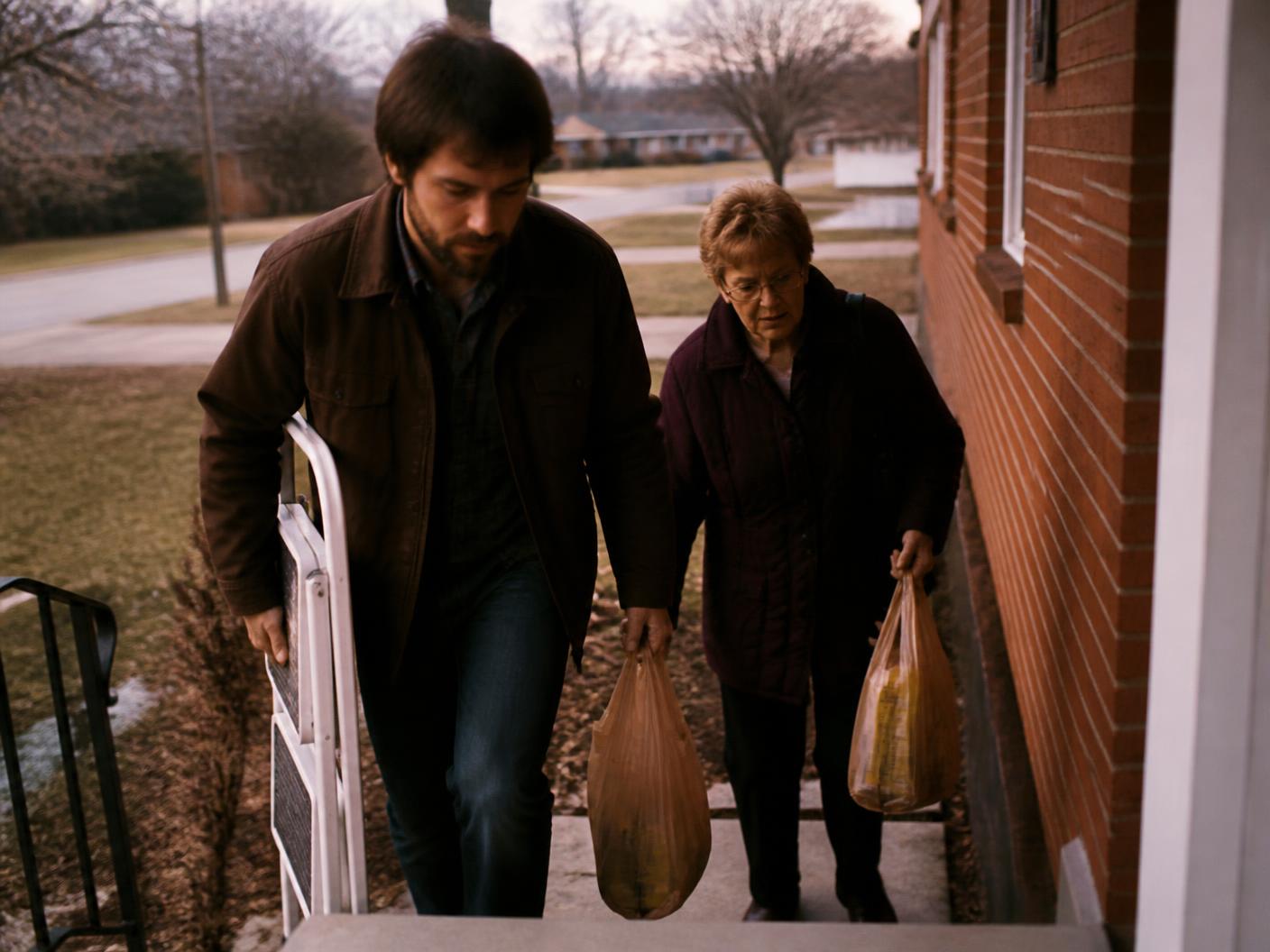 A neighbor delivering a box of fresh groceries to a smiling older woman at her door